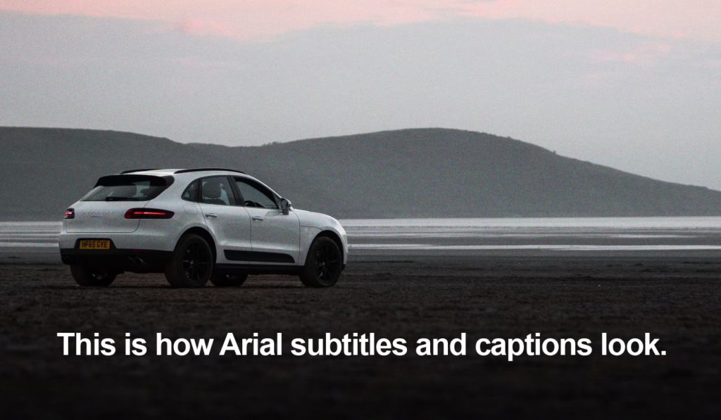 A white Porsche Macan parked on a beach with hills in the background at sunset. Text overlay reads: "This is how Arial subtitles and captions look.